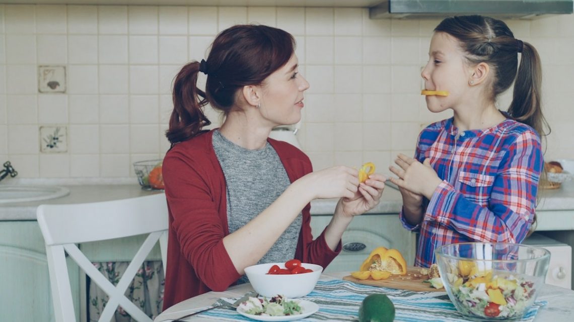 Joyful mother and cheerful daughter have fun grimacing silly with vegetables while cooking in the kitchen at home. Family, cook, and people concept