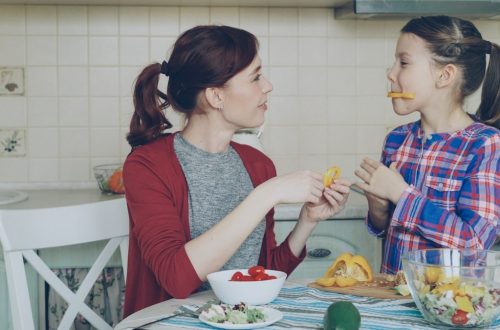 Joyful mother and cheerful daughter have fun grimacing silly with vegetables while cooking in the kitchen at home. Family, cook, and people concept