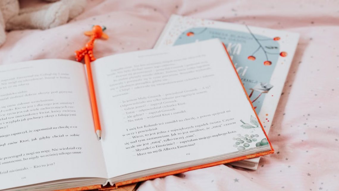 An open book with an orange pencil resting inside, on a star-patterned pink bedsheet.
