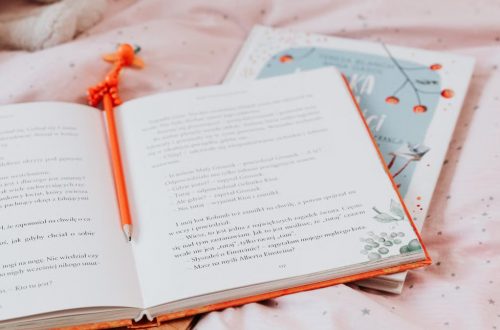 An open book with an orange pencil resting inside, on a star-patterned pink bedsheet.