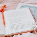 An open book with an orange pencil resting inside, on a star-patterned pink bedsheet.